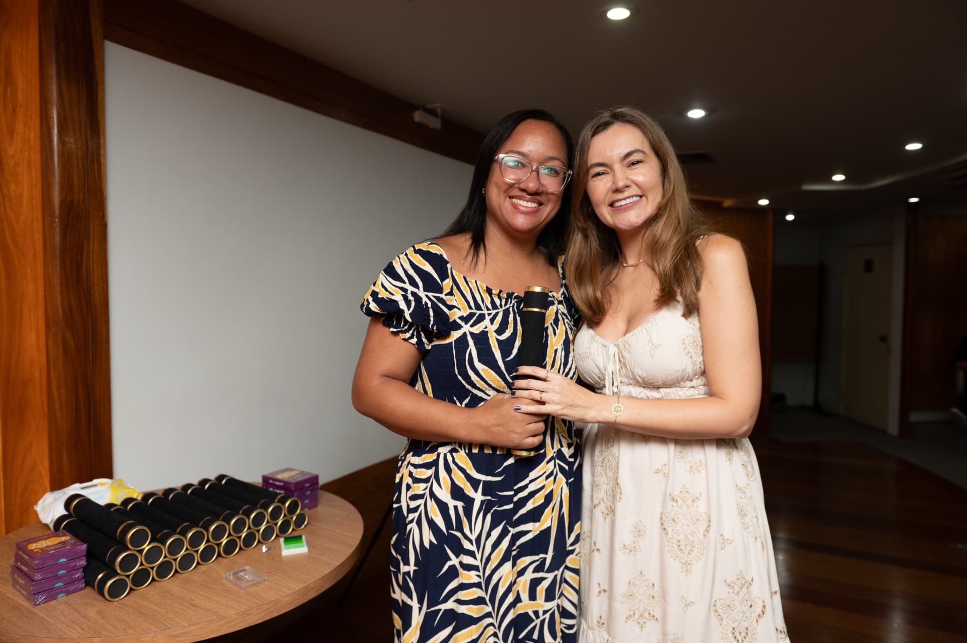 Turma 01 da Pós 2026 — aluna de vestido estampado com o canudo, ao lado da mesa de diplomas